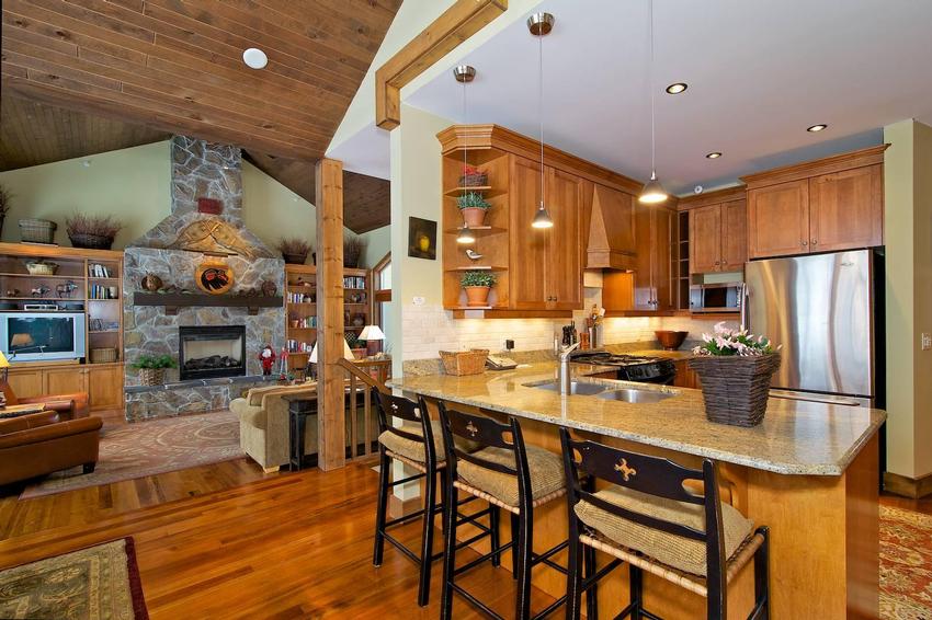 The open concept dining and kitchen area with the living room in the background, with classic ski chalet styling of a stone fireplace and wooden accents, in a South Point luxury townhome at Big White Ski Resort, managed by Luxury Mountain Vacation Rentals.