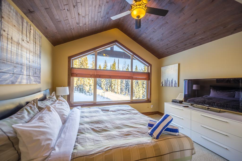 A cozy, comfortable king bedroom with a pointed accent large window and panelled ceiling in a luxury Big White vacation rental.