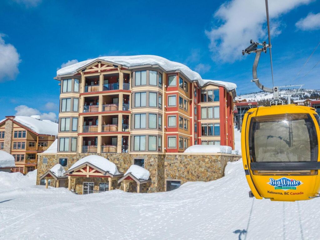One of the Big White Ski Resort Gondolas travelling down the mountain on a sunny day with powdery snow on the ground and The Timbers building in the background in Happy Valley.