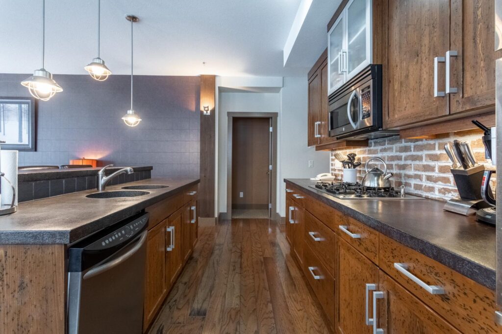 A luxury modern kichen with darak granite countertops, dark wooden cabinets and hardwood floors, and stainless steel appliances with white walls and bright natural light, managed by Luxury Mountain Vacation Rentals at Big White Ski Resort.