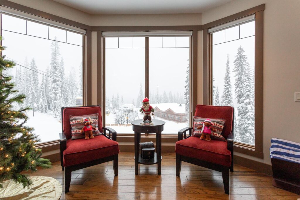 Two red chairs sit in a large window bay with a Christmas tree on the left hand side, and a snowy background through the windows while at Big White Ski Resort.