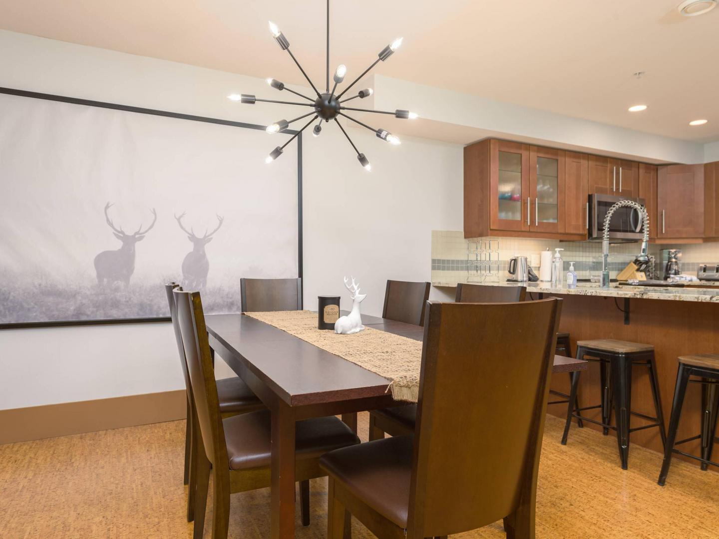 Raven 304's open concept dining space with the gourmet kitchen in the background, a large wooden table and a modern chandelier above, managed by Luxury Mountain Vacation Rentals at Big White Ski Resort