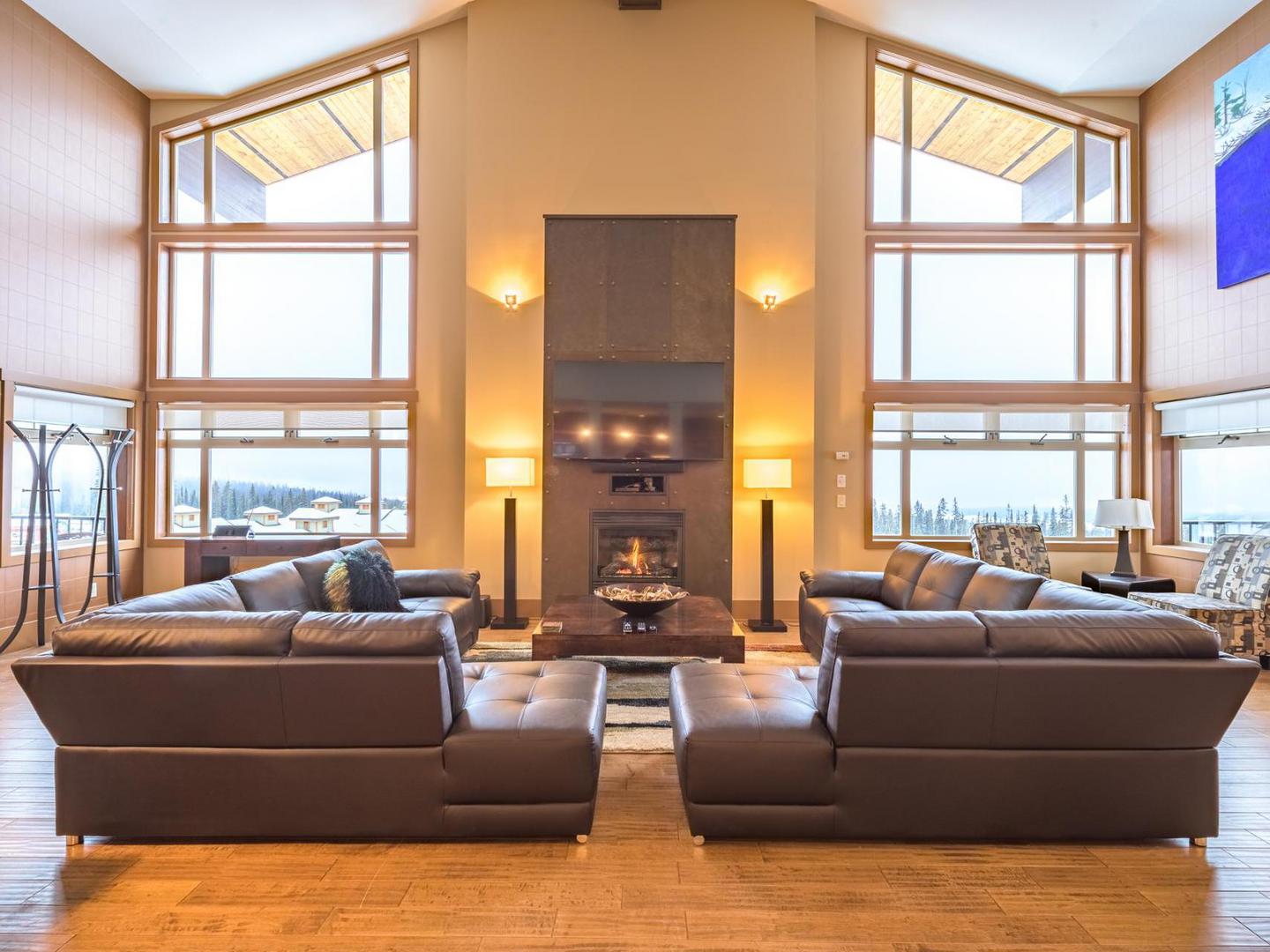A living room with large leather corner couches, high ceilings and floor to ceiling windows in one of Luxury Mountain Vacation Rentals' units at Big White Ski Resort.