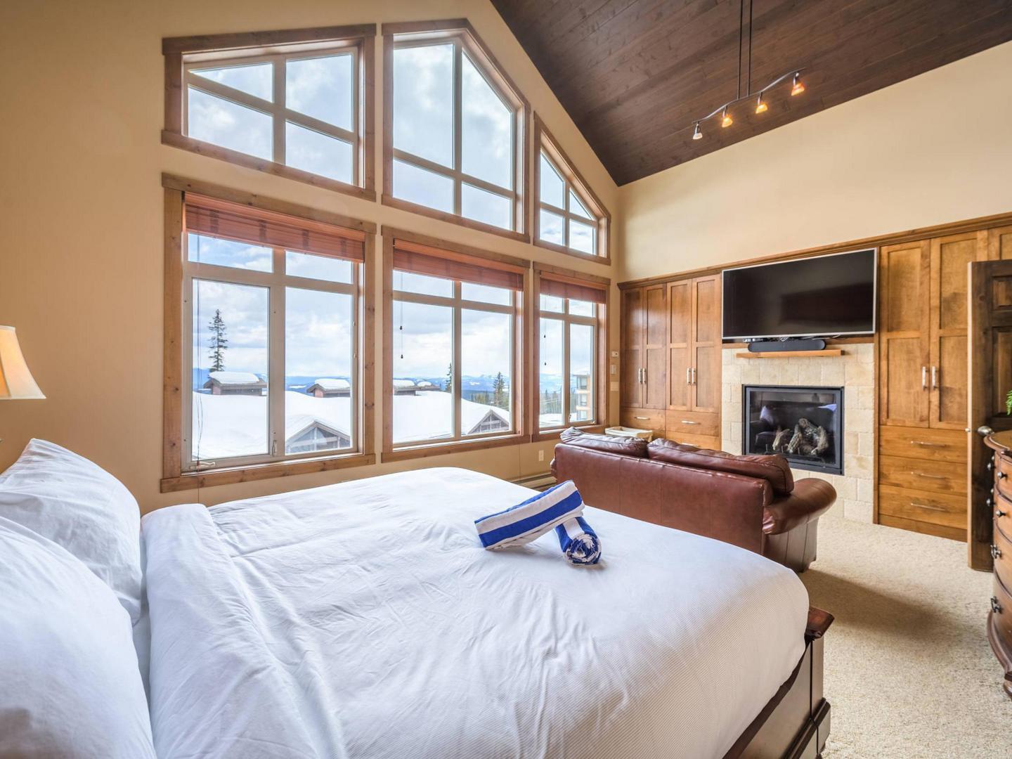South Point 26 penthouse condo rental's master bedroom with a large cozy bed, a fireplace and floor to ceiling windows, managed by Luxury Mountain Vacation Rentals at Big White Ski Resort.