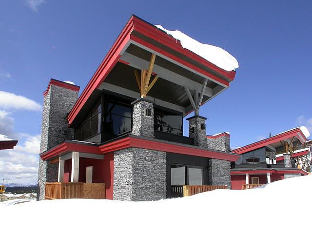 The exterior of a modern ski chalet with red accents and stone pillars, with a snowy trial going by the front on a sunny day, one of the Edge properties managed by Luxury Mountain Vacation Rentals at Big White Ski Resort.