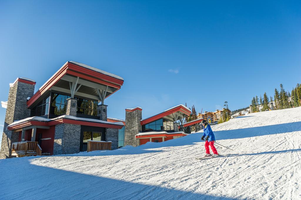 A lone skier in a blue jacket and red pants skis down a ski hill past one of the Edge ski chalets, a luxury modern ski chalet managed by Luxury Mountain Vacation Rentals and located at Big White Ski Resort.