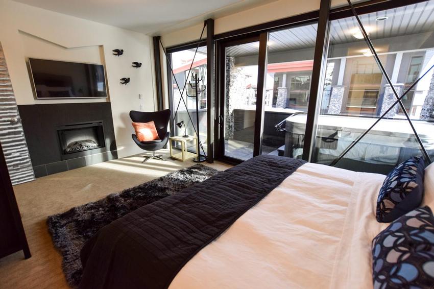 A cozy, large master bedroom with a large bed, bright natural light from the large windows and balcony glass door, seating and a fireplace in a vacation rental by Luxury Mountain Vacation Rentals at Big White Ski Resort.