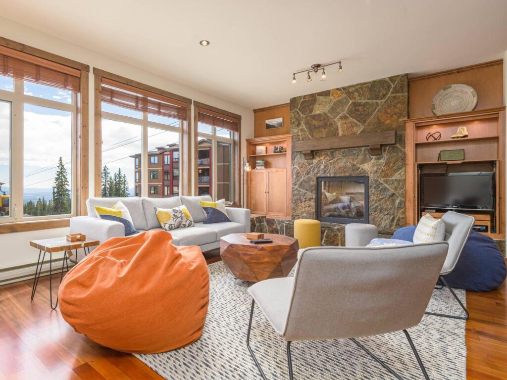 A cozy, modern-style living room with hardwood floors, stone fireplace and bright, large windows in a Luxury Mountain Vacation Rentals vacation rental at Big White Ski Resort.