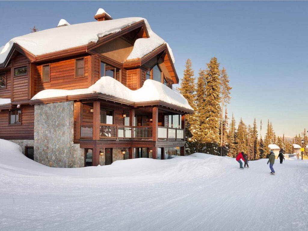 The outside of White Caviar, a luxurious ski mountain vacation rental with wooden and stone accents and covered in snow, with skiers going by on a snowy sunny day, one of Luxury Mountain Vacation Rentals' units on Big White Ski Resort.