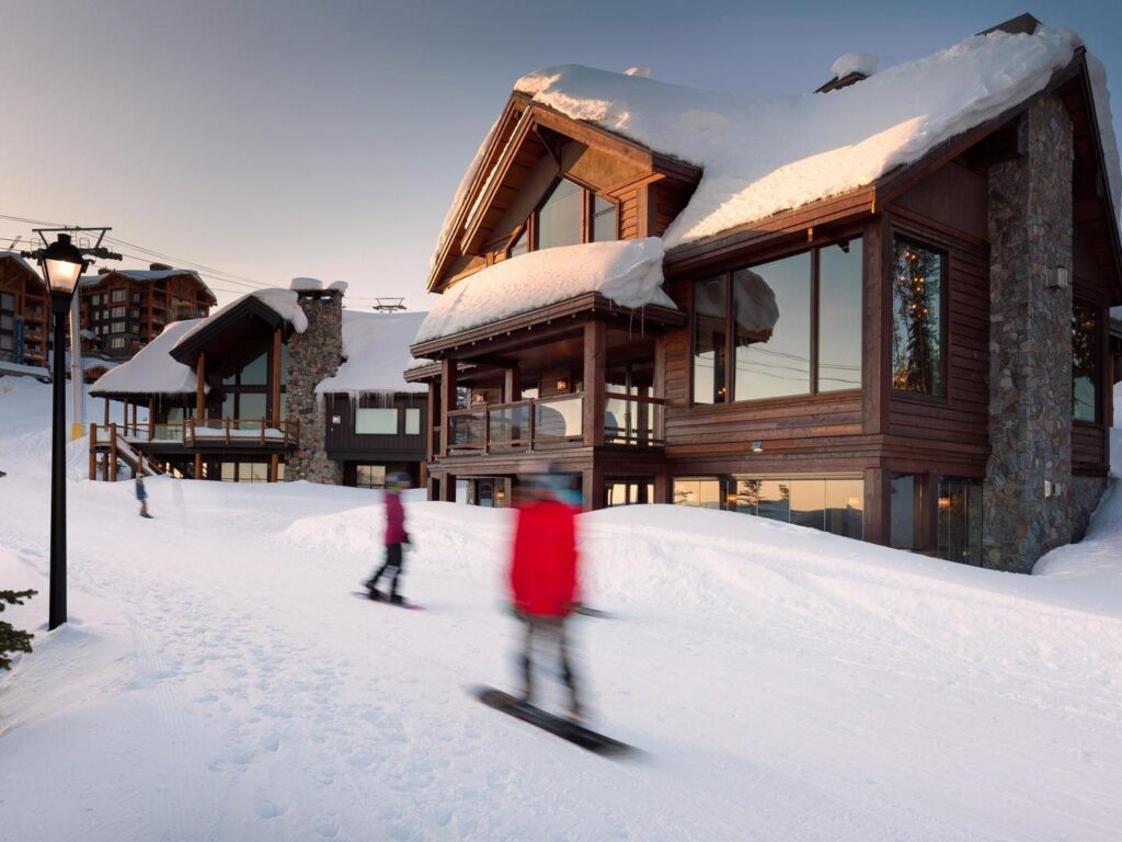 Snowboarders zoom past a luxury ski chalet on a snowy day near sunset at Big White.