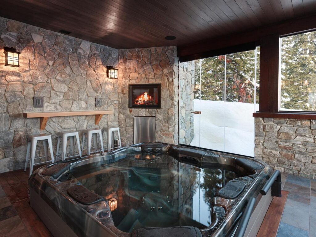 A reflective hot tub on a covered patio with stone walls and wooden stools in the background on a snowy day in a luxury rental from Luxury Mountain Vacation Rentals at Big White Ski Resort 