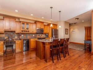 A gourmet kitchen with wooden cabinets and stone countertops, with hardwood flooring and a bar with benches at the kitchen island, makes up a gourmet kitchen in one of Luxury Mountain Vacation Rentals' suites at Big White Ski Resort
