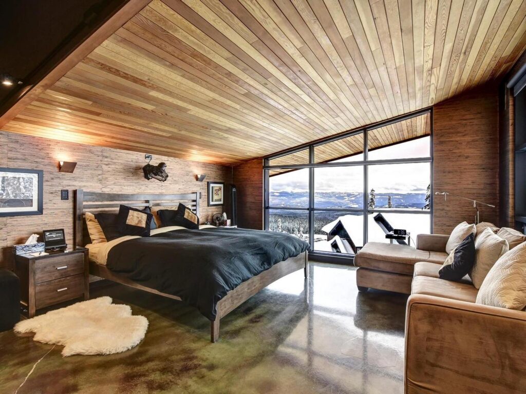 A luxury bedroom with a wood panelled, lofted and slanted ceiling, with large floor to ceiling windows, warm lighting and luxury touches in a Luxury Mountain Vacation Rentals suite at Big White Ski Resort