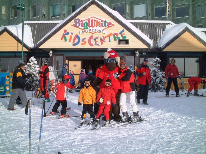 A family with their kids all in skis and ski gear stand with the Big White mascot, the moose, in front of the Big White Kids Centre at Big White Ski Centre, a perfect activity for kids while on a family vacation.