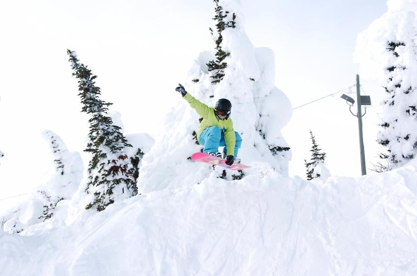 A snowboarder in a lime green jacket and bright blue pants makes a jump over a snowbank along a ski trail at Big White Ski Resort.