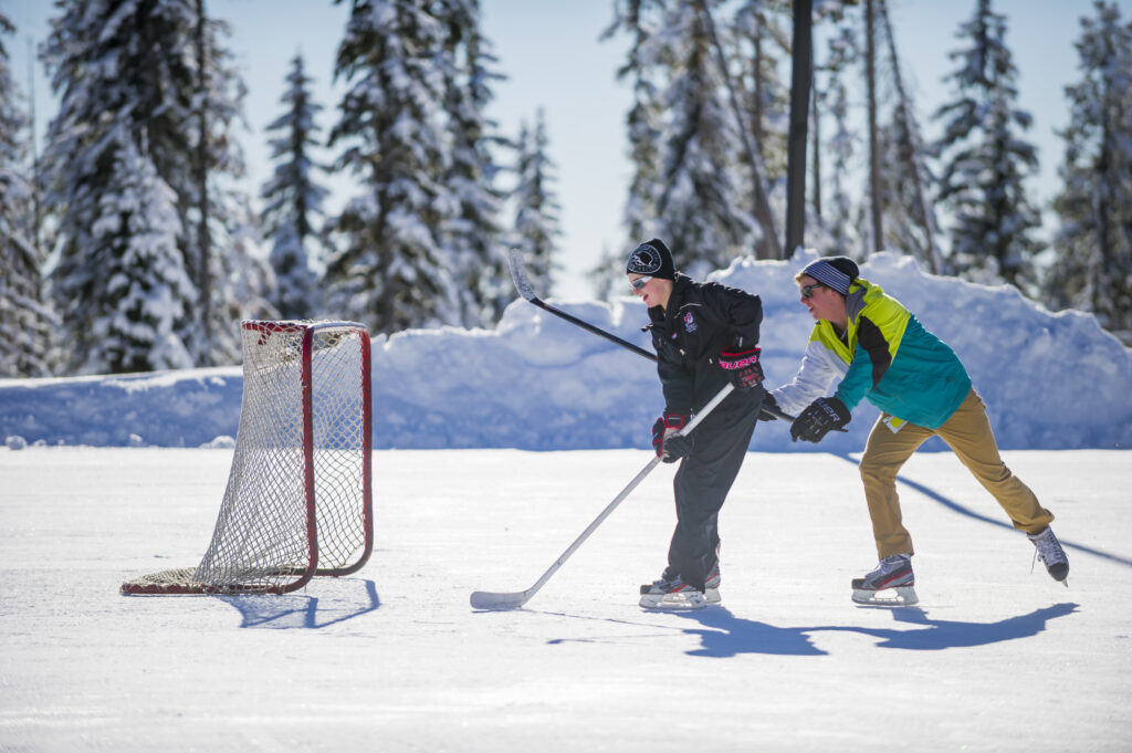 Two children with hockey sticks are playing ice hockey and shooting on a small net on an outdoor ice rink at Big White Ski Resort