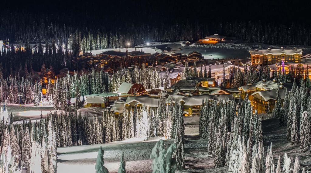 A faraway view of Big White Ski Resort at night, covered in fluffy white snow and surrounded by snow covered evergreen trees, along with splashes of warm light from the buildings in the Village Centre.