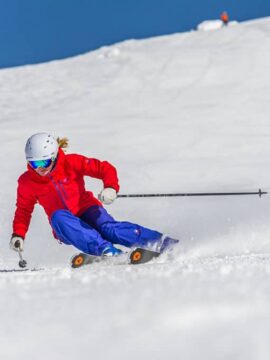 A skier dressed in red and blue skis down a snowy trail on the side of the mountain at Big White Ski Resort, with bright blue skies and freshly packed, champagne powder snow.