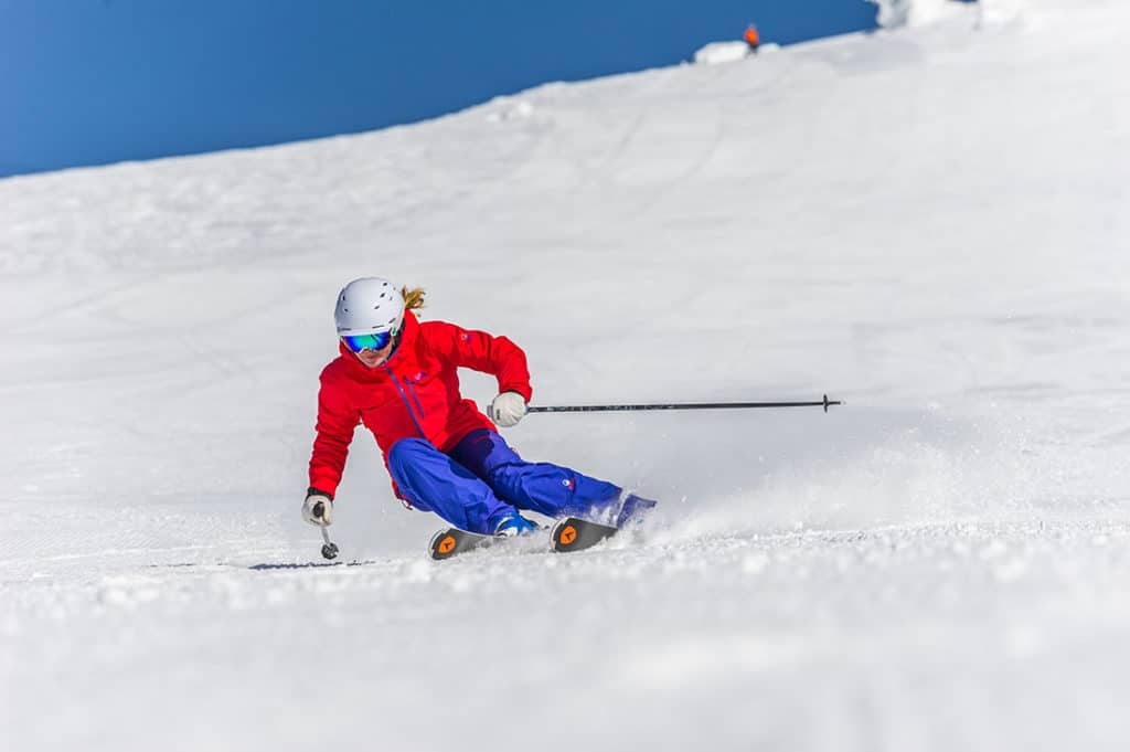 A skier dressed in red and blue skis down a snowy trail on the side of the mountain at Big White Ski Resort, with bright blue skies and freshly packed, champagne powder snow.