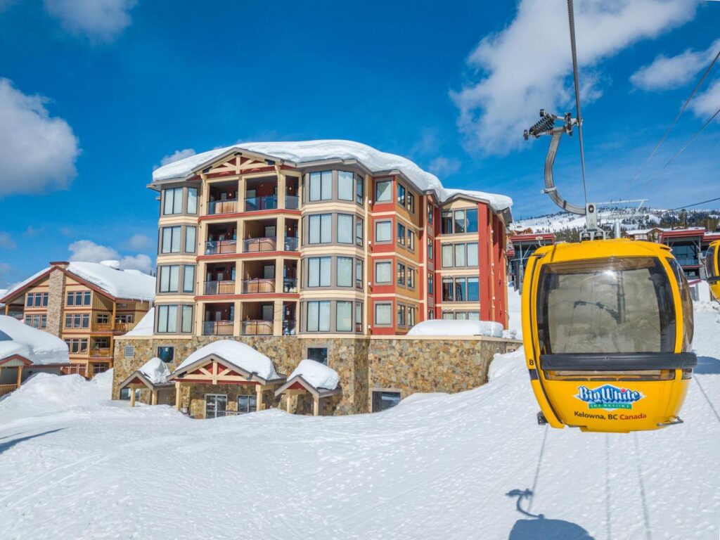 A Big White gondola travelling down the mountain past a vacation rental building on a sunny and snowy day in Happy Valley.