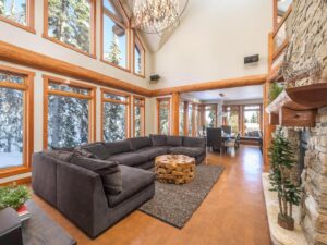 A large grey sectional couch in a spacious living room with a stone fireplace to the right, and floor to ceiling windows on the left, and the dining room in the back in one of Luxury Mountain Vacation Rentals' suites.