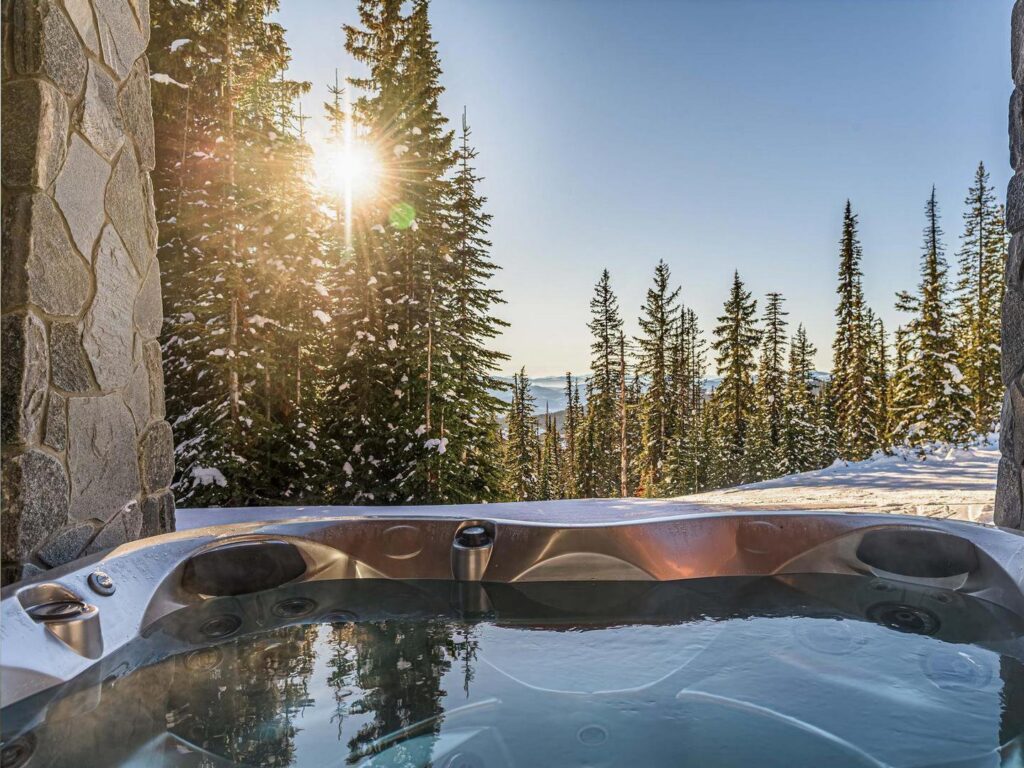 A relaxing hot tub overlooks a bright sunny sky and evergreen trees at Big White Ski Resort, the perfect spot for a mountain wellness retreat.