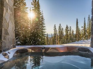 A reflective, still hot tub on a private patio in one of Luxury Mountain Vacation Rentals' luxury suites, with a view of the setting sun over evergreen trees and snow on Big White.