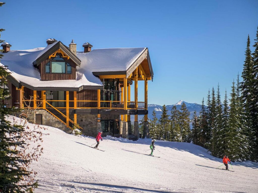 Three people ski past a luxury ski chalet on a sunny day at Big White.