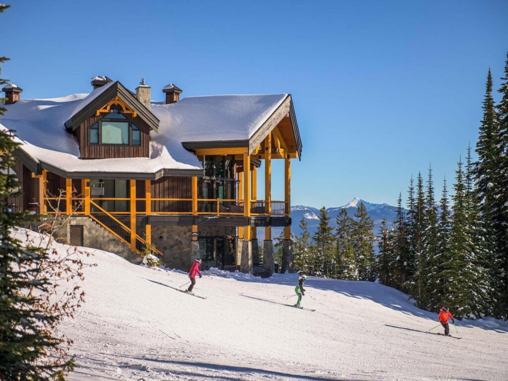 Three skiers ski down a snowy hill past a large luxury rental building at Big White Ski Resort with bright blue skies above.