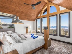 A coxy, large bed with white blankets and grey pillows in a bedroom with soft carpeting, wood-panelled ceiling and a huge, floor to ceiling window overlooking Big White Ski Resort in one of Luxury Mountain Vacation Rentals' suites.