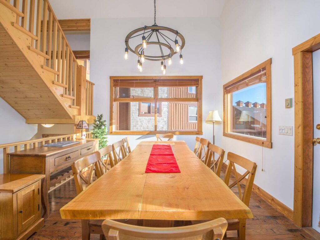 A bright, open dining room with a large dining table, stairs on the left, large windows and a modern chandelier overhead in a luxury vacation rental from Luxury Mountain Vacation Rentals at Big White Ski Resort.