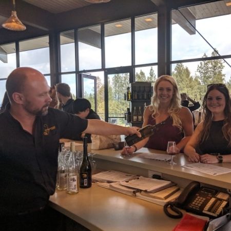 A winery bartender pours a glass of wine for a pair of smiling women standing at the winery bar on their wine tasting tour with Discover Okanagan Tours.