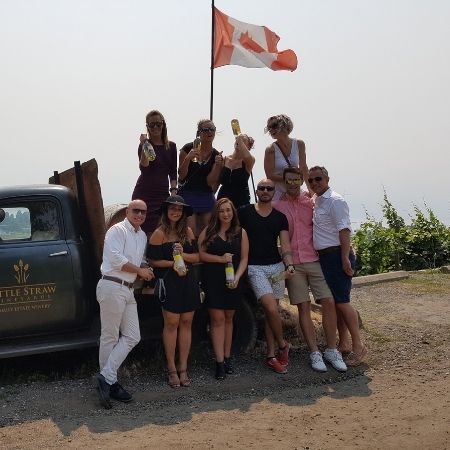 A group of friends standing in front of or on a wine truck with a canadian flag, with all of them holding a bottle of wine they got on their wine tour with Discover Okanagan Tours.