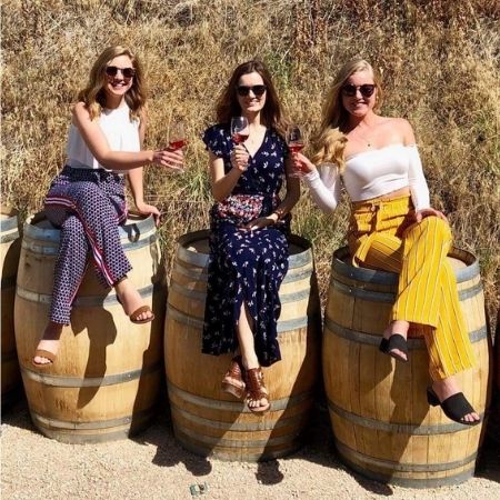 A trio of ladies dressed in bright summery clothes, sitting on top of wine barrels and saying cheers with their glasses of wine on a Discover Okanagan Tours wine tour.