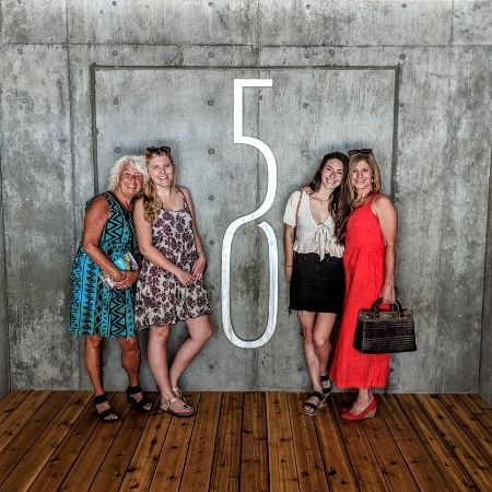 A group of four ladies in dresses standing in front of a logo on a contemporary concrete wall for a winery, posing for their wine tour photo with Discover Okangan Tours