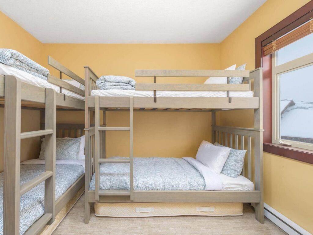 A bedroom in one of the Aspens townhomes, with yellow walls, bright windows and two bunkbeds, managed by Luxury Mountain Vacation Rentals at Big White Ski Resort.