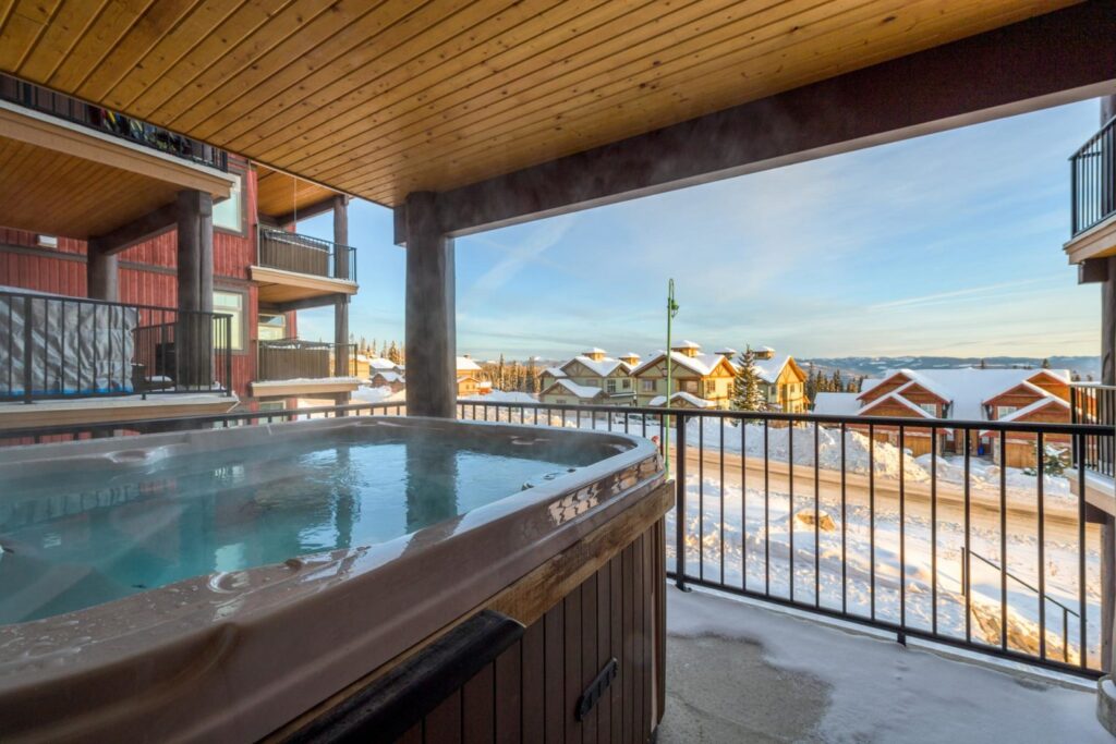 Pet-friendly Raven Unit 205's private deck with a cozy private hot tub, with views looking over Big White Ski Resort, on a sunny, snowy day.