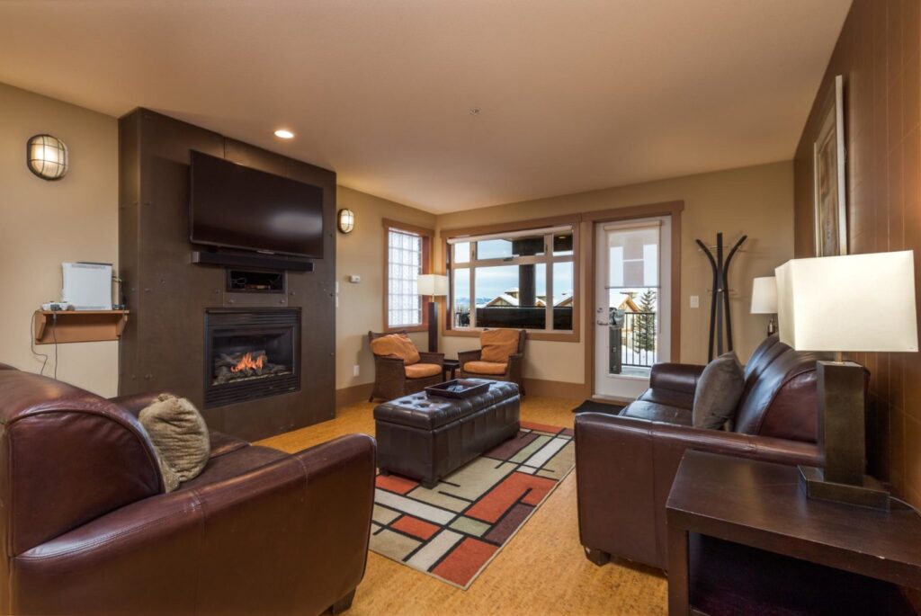 Pet-friendly Raven Unit 205's spacious and comfortable living area, with a gas fireplace, bright windows, two couches and multiple seating arrangements, and a red and square patterned carpet on wooden floors.