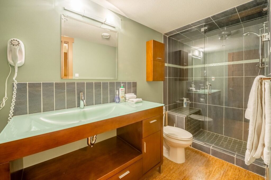 Pet-friendly Raven Unit 209's luxury, spa-like bathroom, with tinted glass counters, chestnut coloured cabinets, grey tiles and wooden floors with a steam shower, up on Big White Ski Resort