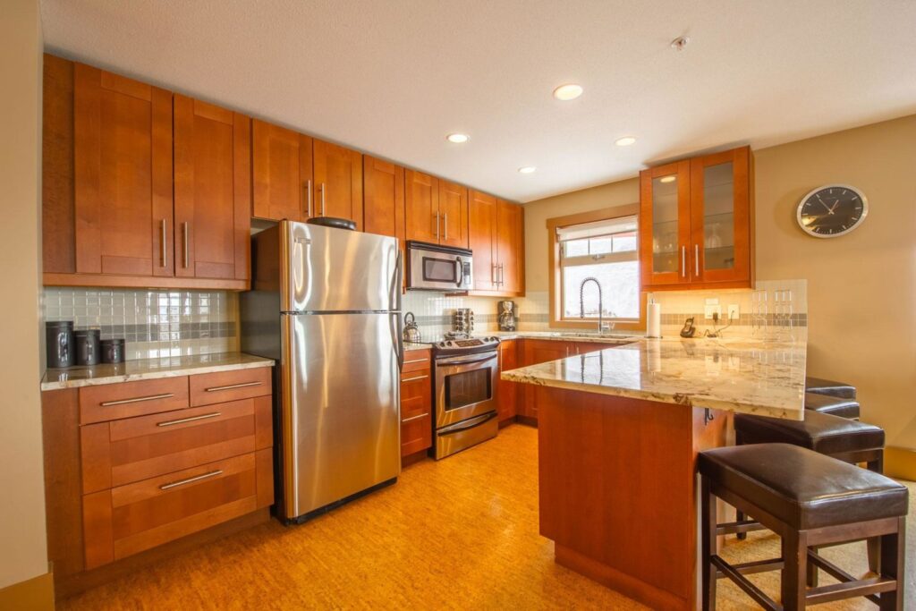 Pet-friendly Raven Unit 209's gourmet luxury kitchen, with chestnut stained wooden cabinets, stainless steel appliances, wooden floors, and comfortable bar stools at the marbled countertop along with bright windows.