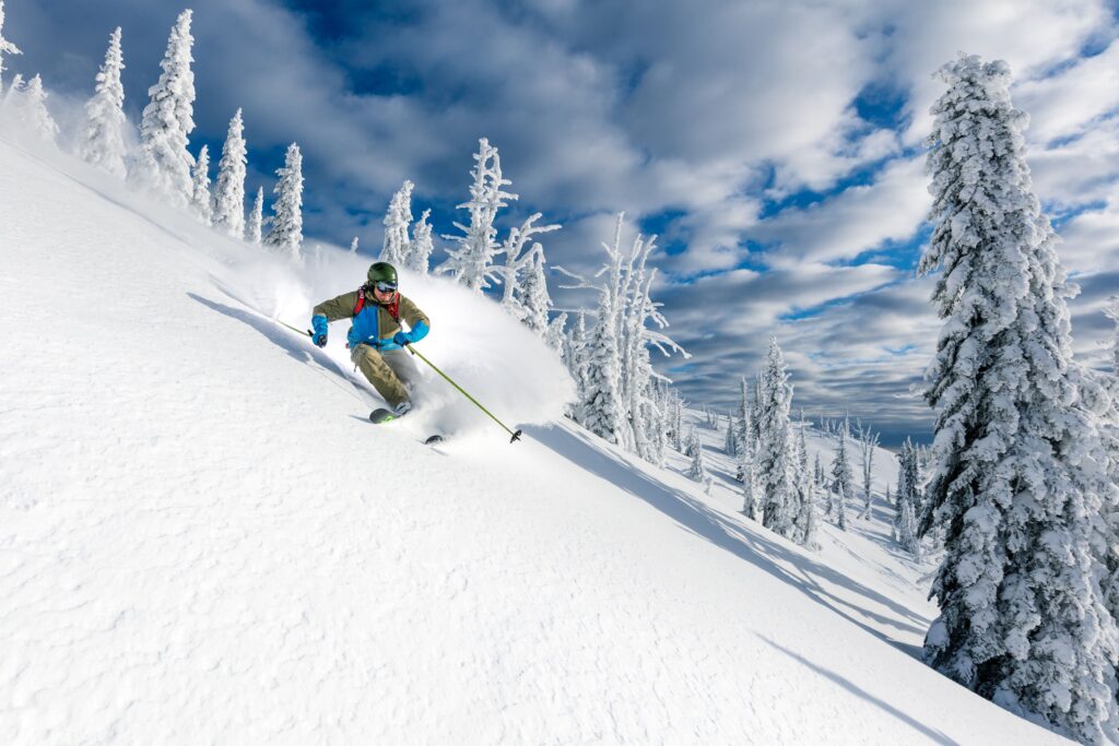 A skier cat skiing down one of Big Red Cats' trails, which is covered in powdery white snow on a blue sky day with fluffy white clouds and bright sun.