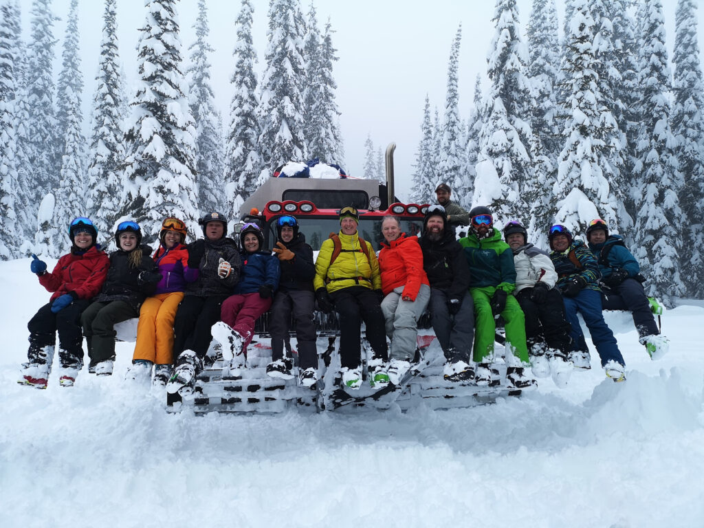 A group of cat skiers sitting on a Cat machine and posing for the photo, in snowy terrain with snow covered trees, on a ski excursion with K3 Cat Ski