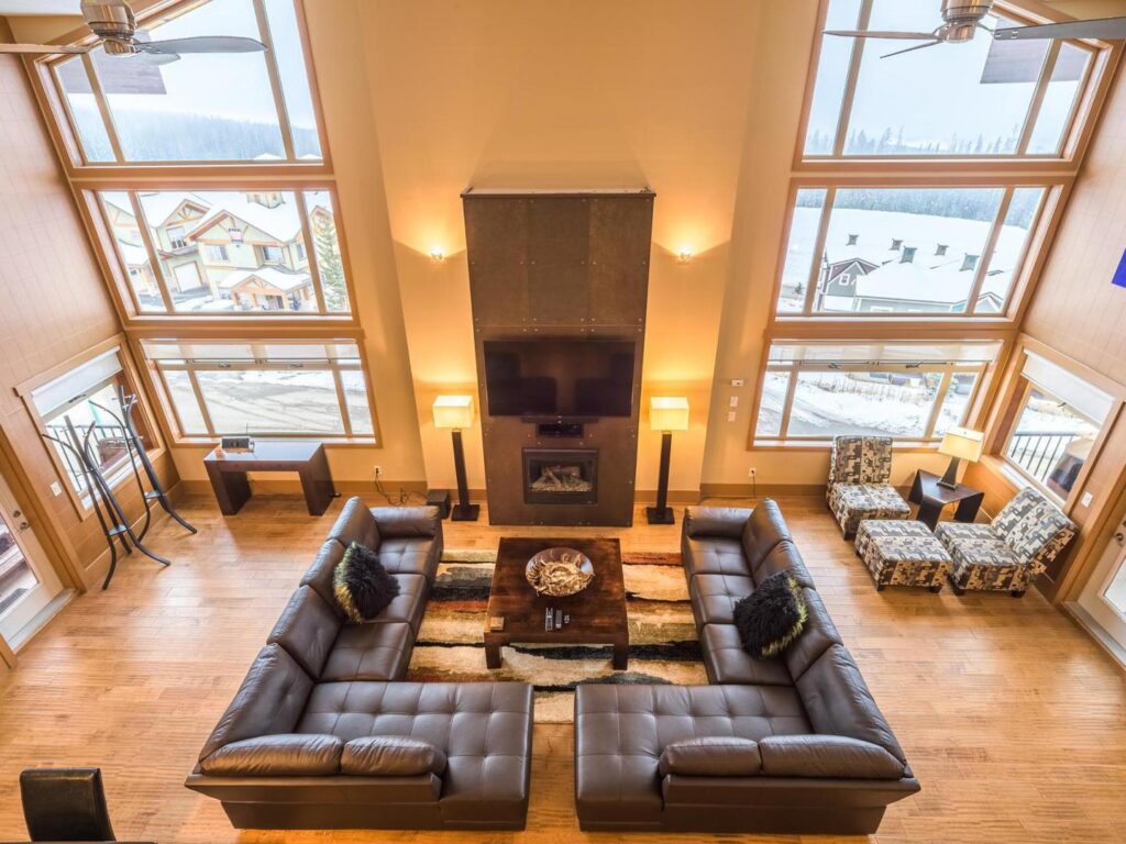 An overhead view of the Great Room in one of Luxury Mountain Vacation Rentals' luxury rentals at Big White Ski Resort, which has two full corner couches, large floor to ceiling windows overlooking a snowy landscape, wooden floors and bright lights.
