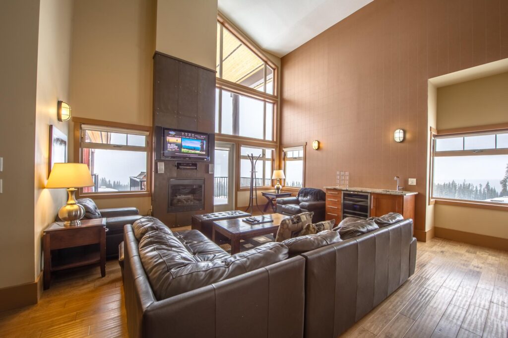 A cozy living room with a full corner leather couch, large TV over a warm gas fireplace and plenty of coffee tables and bright lights, along with large windows overlooking the beautiful snowy Big White Ski Resort, in one of LMVR's luxury rental accommodations.