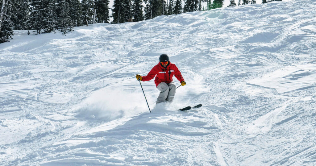 A skier in a red jacket skis down a snowy slope with tall evergreen trees in the background while at Big White Ski Resort while going skiing in January.
