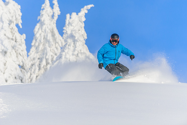 A snowboarder in a blue jacket snowboards down a champagne powder, snowy slope with snow-covered trees in the background and bright blue skies above while at Big White Ski Resort, perfect conditions for snowboarding or skiing in January.