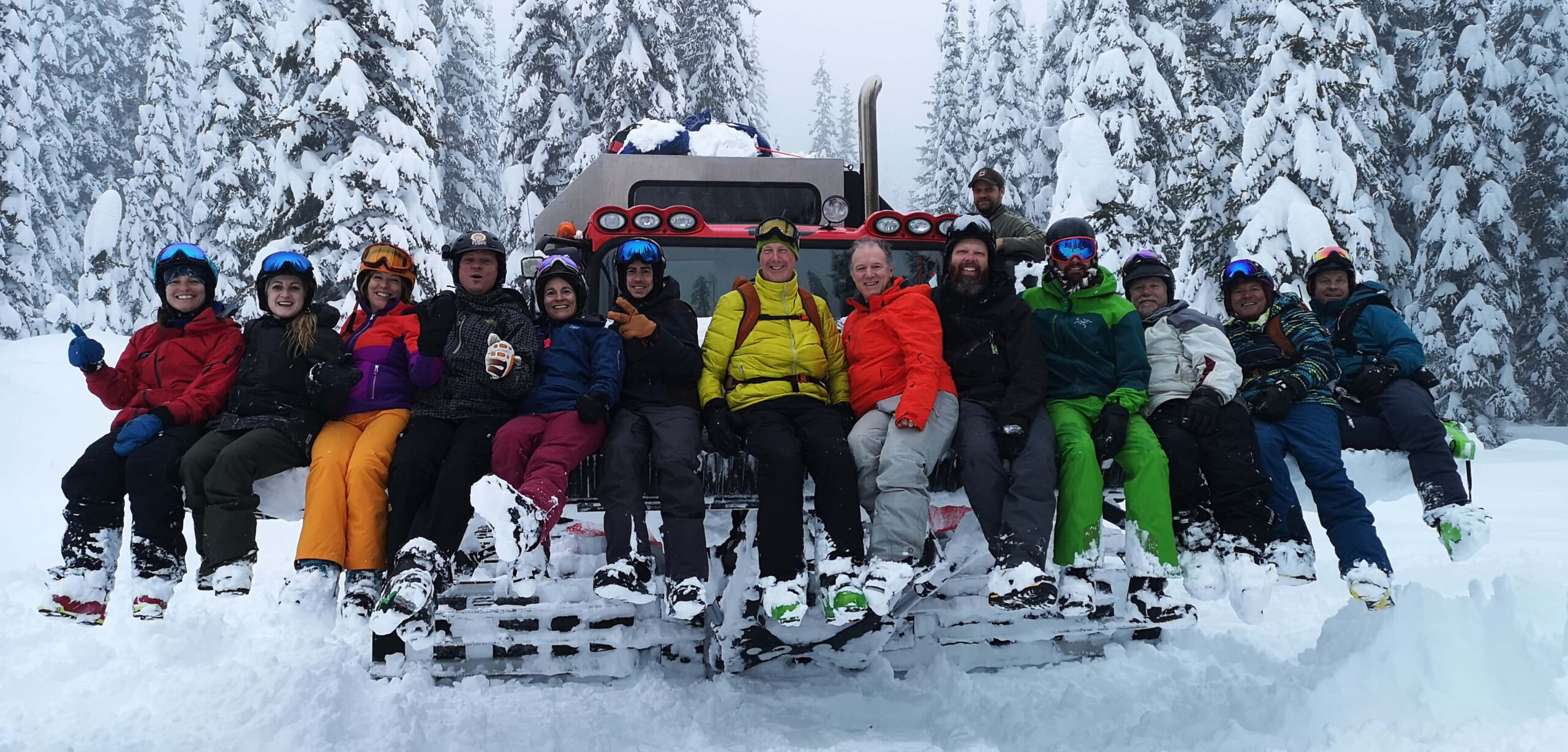 A large group of cat skiers posing by a cat machine for K3 Cat Ski on a snowy day with snow covered trees in the background.