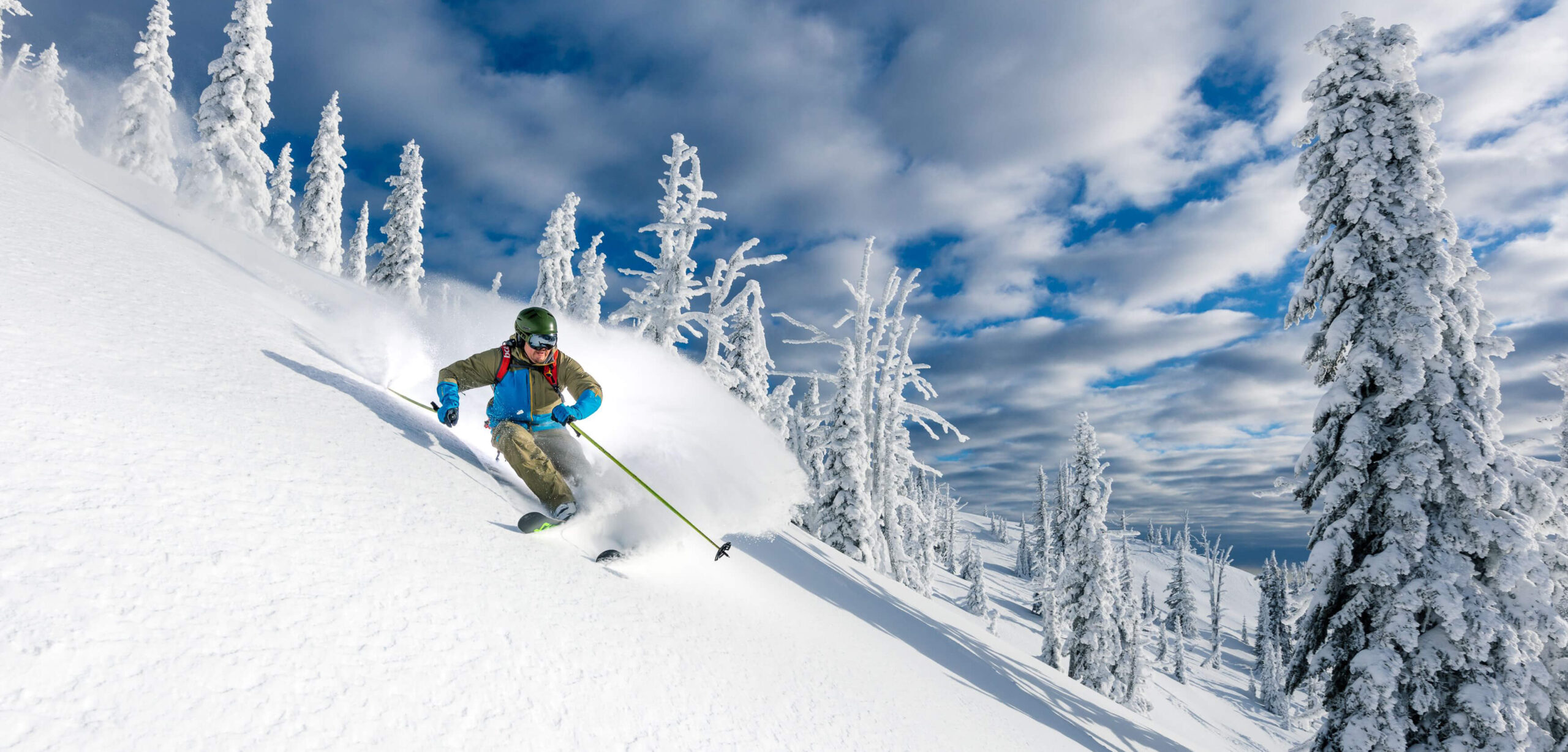 A skier cat skiing down one of Big Red Cats' trails, which is covered in powdery white snow on a blue sky day with fluffy white clouds and bright sun.