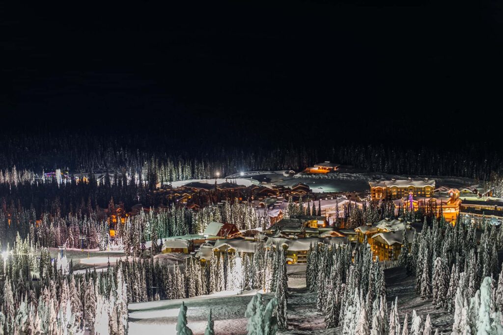 The town of Big White at night, covered in a blanket of snow with warm lighting around the buildings and ski trails at Big White Ski Resort.
