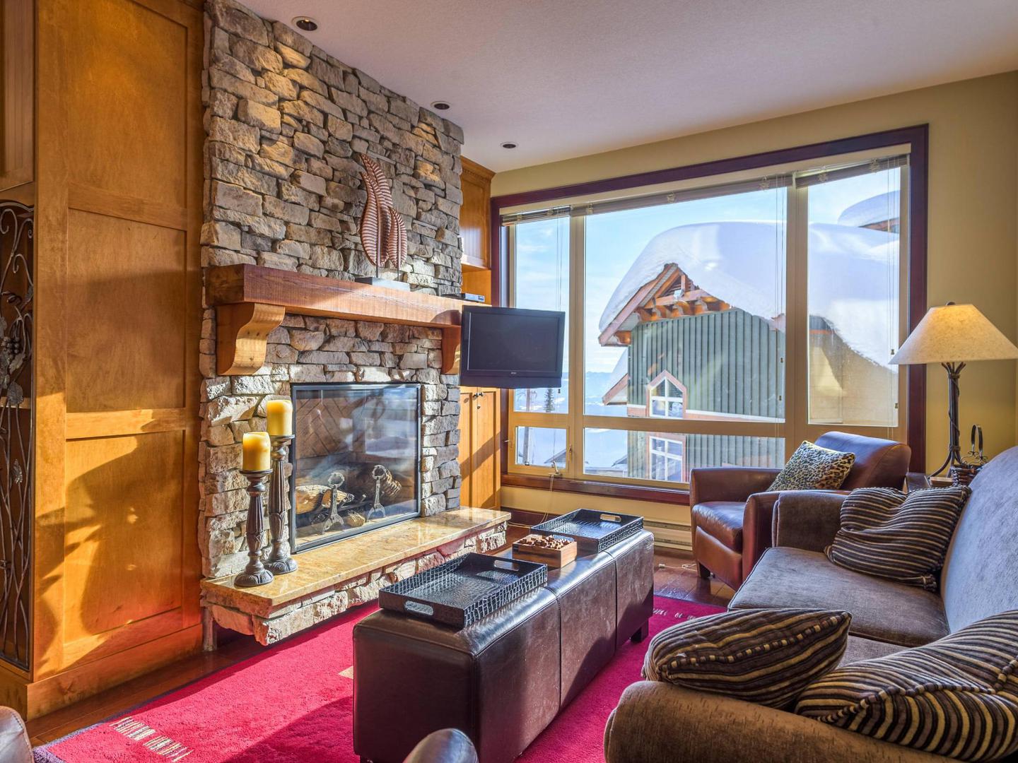 Aspens 201's cozy living room with a comfortable couch, stone fireplace and bright warm light from the floor to ceiling windows, a luxury vacation rental condo managed by Luxury Mountain Vacation Rentals at Big White Ski Resort